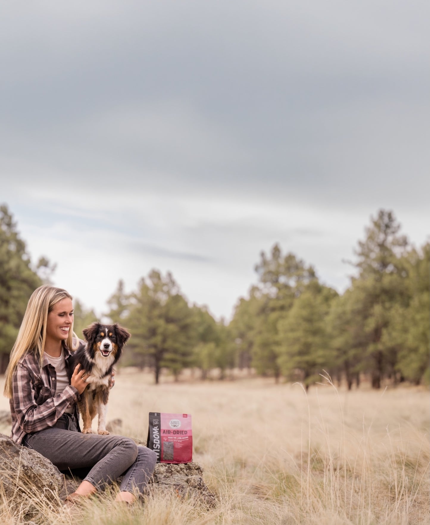 Woman sitting with a dog in a field holding a bag of Wisdom Air-Dried Dog Food.